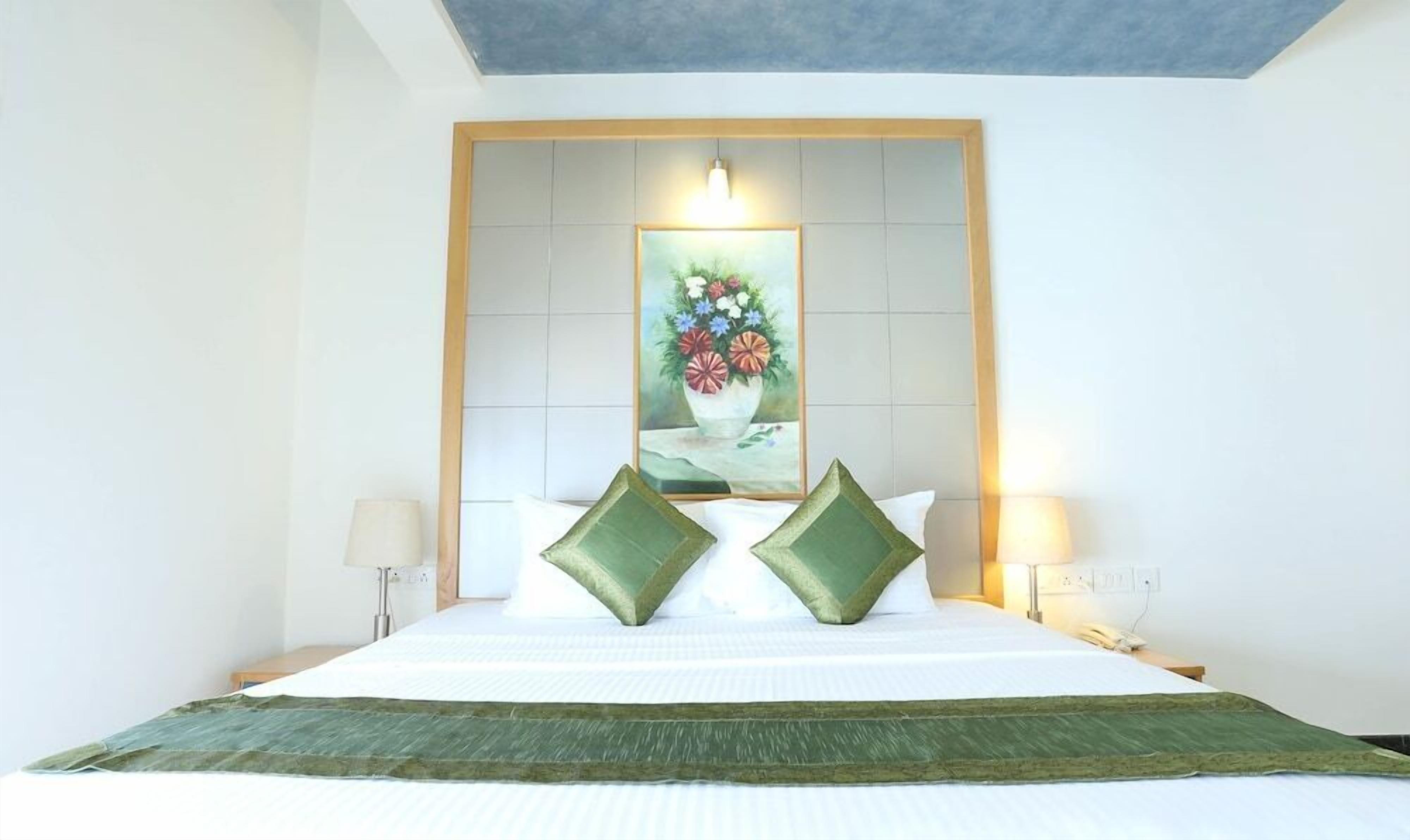 A hotel room with a large bed, two green pillows, a painting of flowers on the headboard, and two bedside lamps.