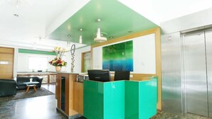 A reception area with a green accent wall, a reception desk, a seating area with a sofa and a small table, and a large window with a view of the sky. - Edha Suites (Bengaluru)