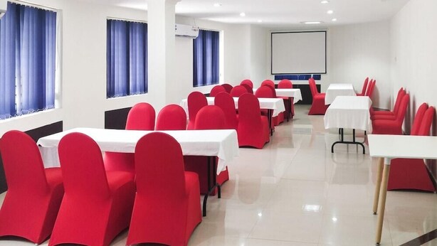 A conference room with red chairs and white tables, large windows with blue curtains, and a projector screen on the wall. - Edha Suites (Bengaluru)