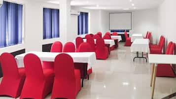 A conference room with red chairs and white tables, large windows with blue curtains, and a projector screen on the wall.