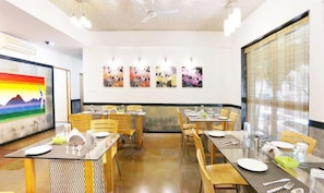 Un comedor con mesas dispuestas para dos, obras de arte en las paredes y un ventanal con persianas.