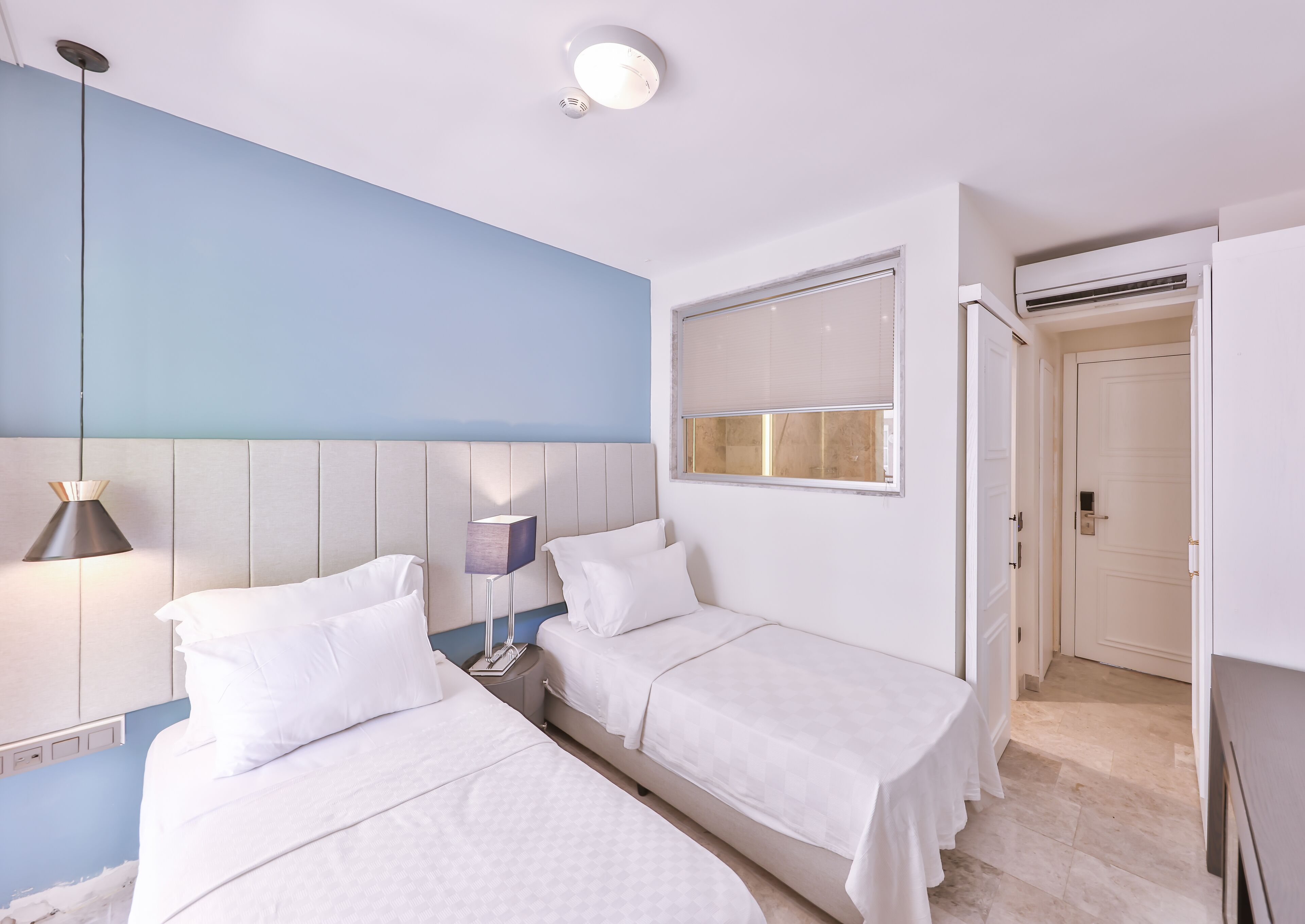 Et hotelværelse med to senge, et natbord, en lampe, et vindue med persienner og en aircondition.
