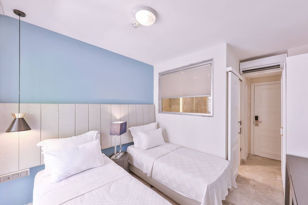Et hotelværelse med to senge, et natbord, en lampe, et vindue med persienner og en aircondition.
