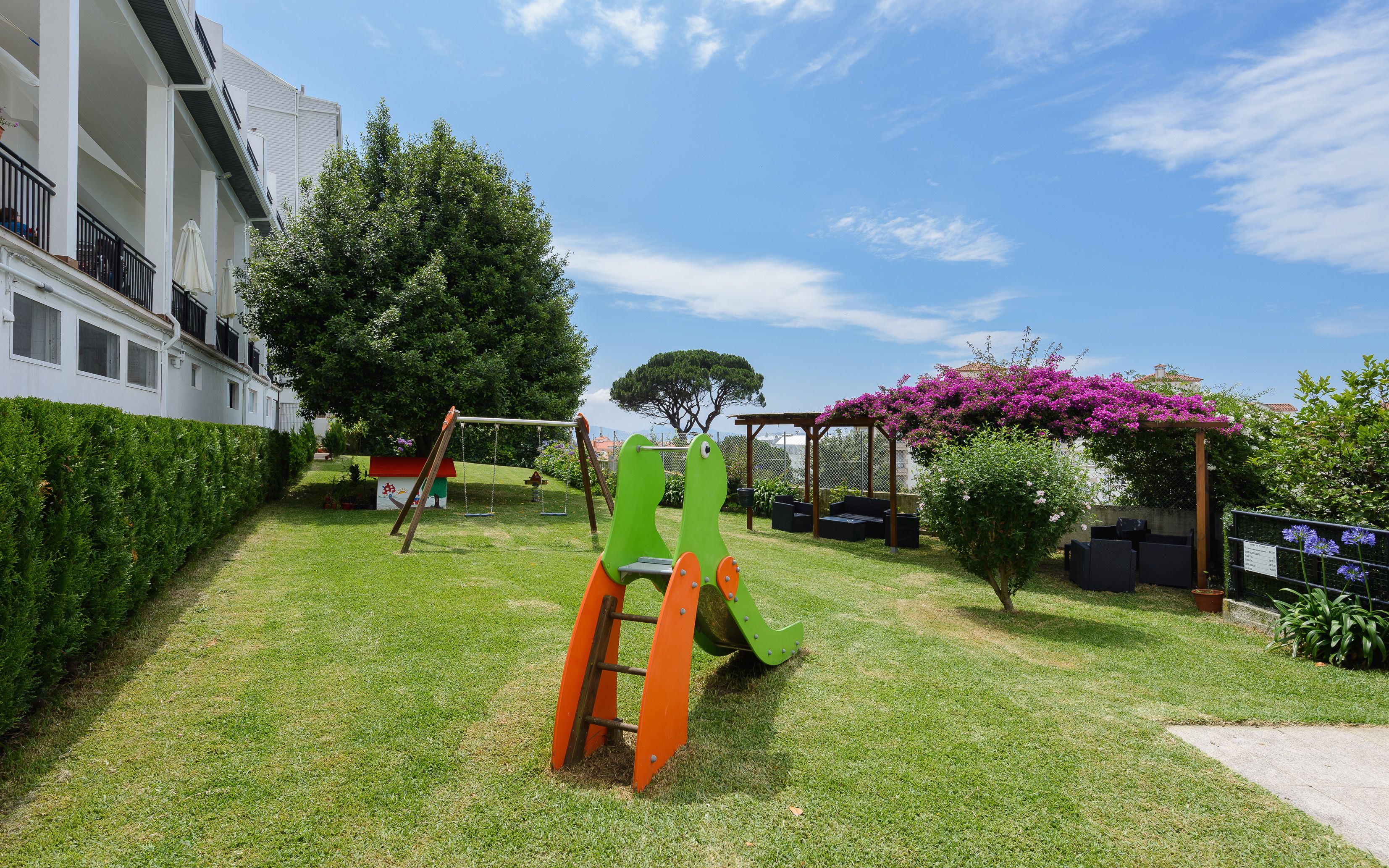 Un espacio exterior cuidado con un columpio, una casita de juegos y una pérgola adornada con plantas en flor.