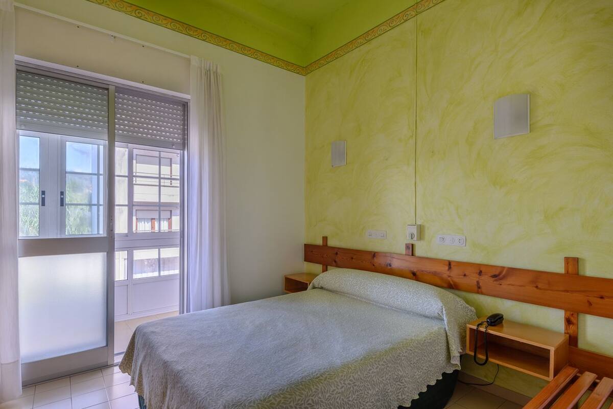 Un dormitorio con cama, cabecera de madera, mesita de noche, teléfono y ventana con cortinas.