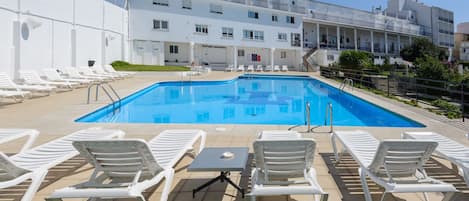 En swimmingpool med hvide liggestole og et bord, omgivet af en bygning med altaner.