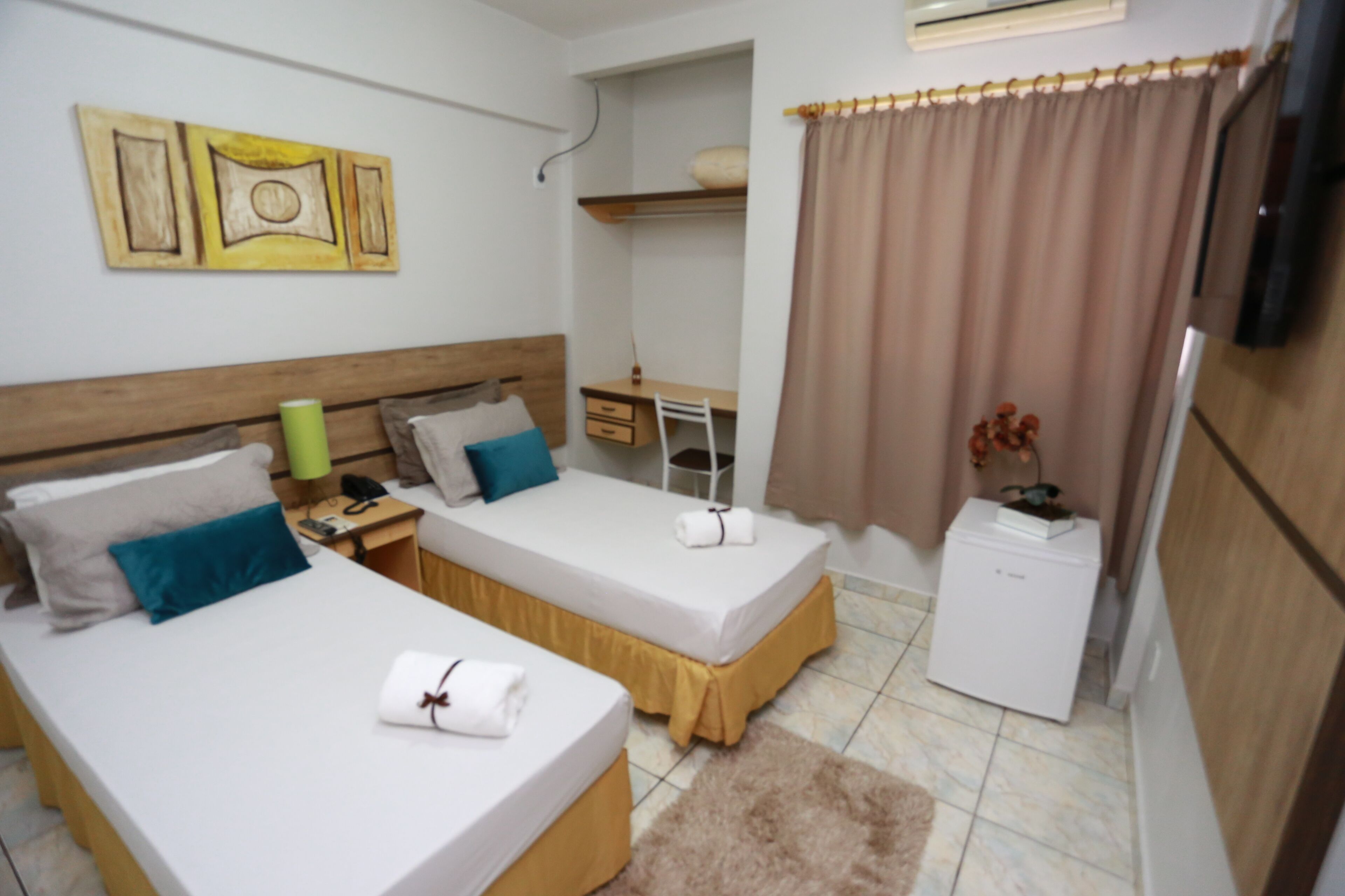 A room with two beds, a desk, a small refrigerator, and a window with curtains.