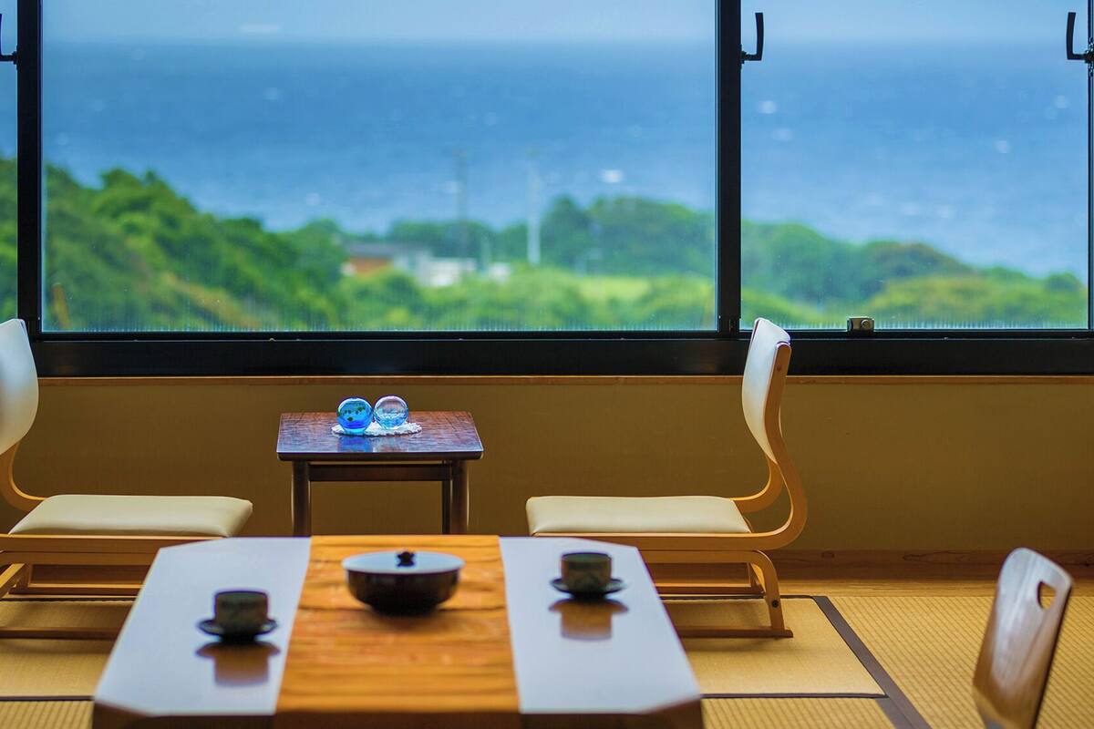 A traditional Japanese room with tatami flooring, a low table with a bowl and chopsticks, two chairs, and a view of the sea through large windows.