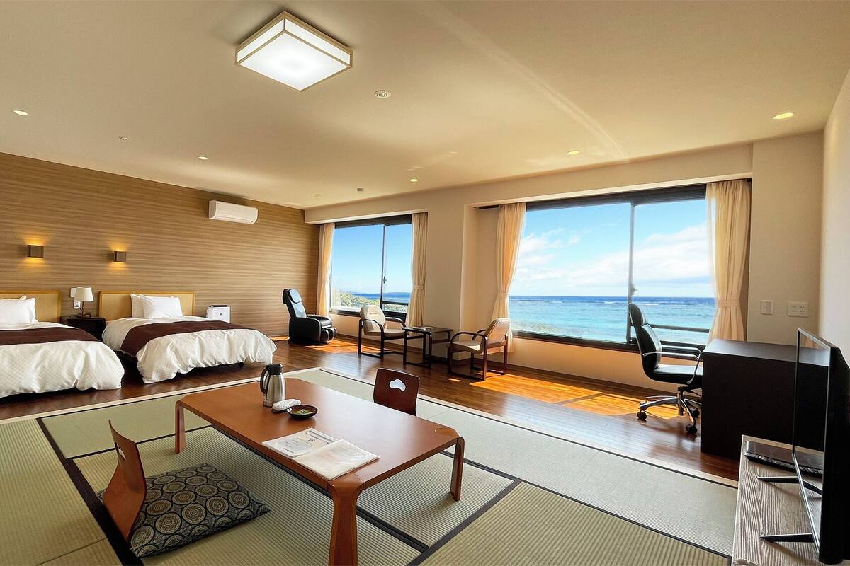 A hotel room with two beds, a desk, a chair, a TV, and a large window with a view of the ocean.