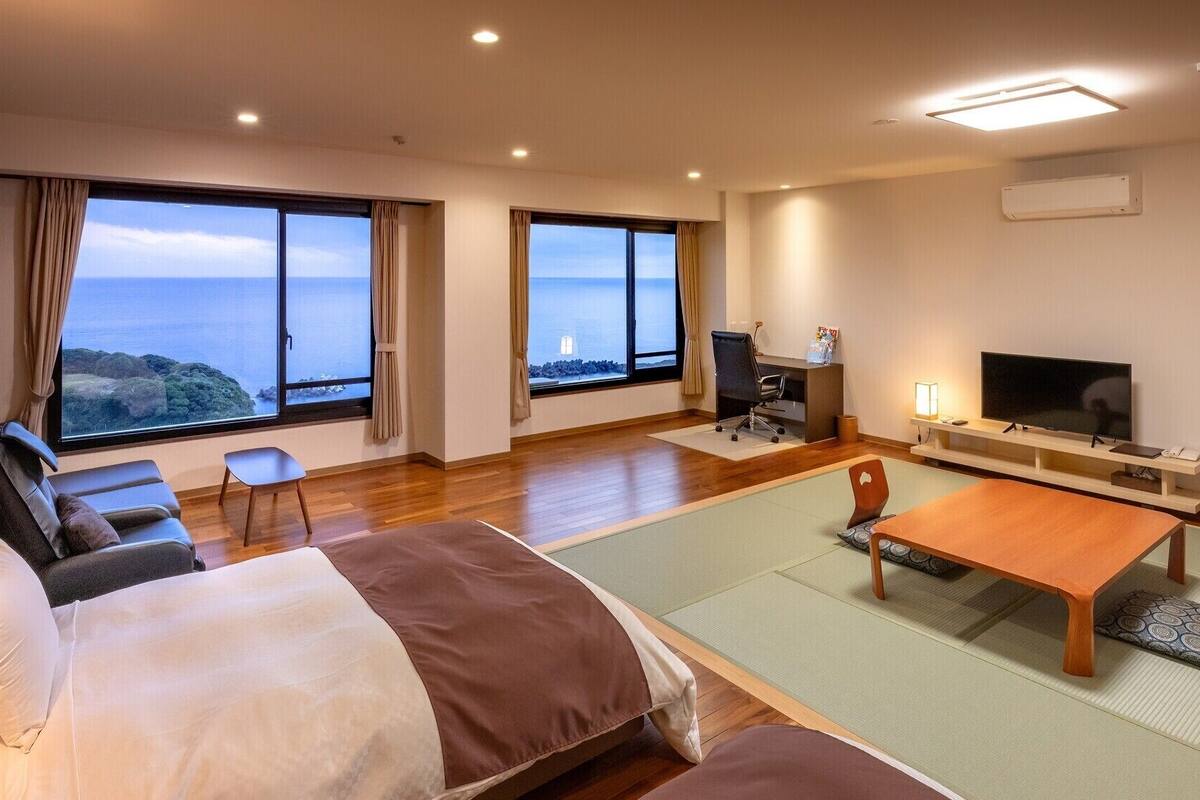 A hotel room with a bed, a desk, a TV, and large windows with a view.