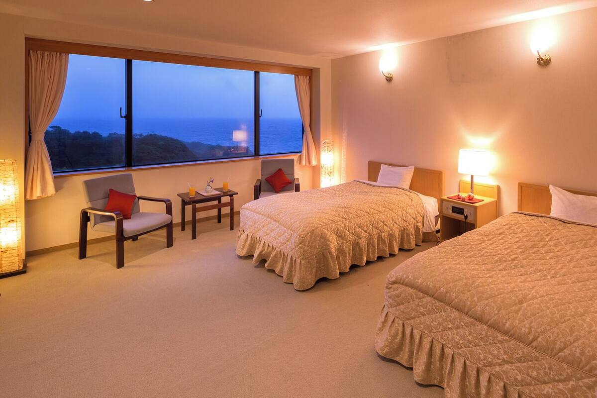A hotel room with two beds, a seating area with a table, and a large window offering a view of the sea.
