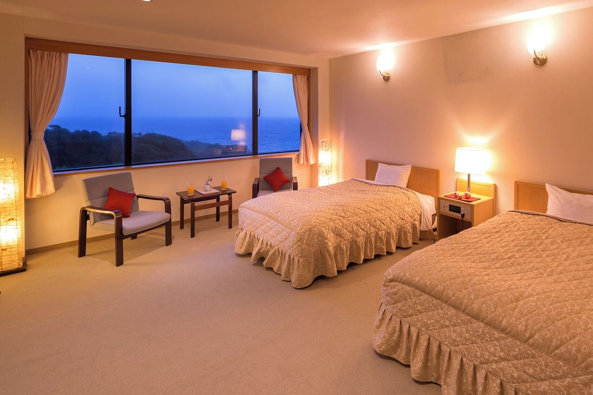Habitación de hotel con dos camas, zona de estar con una mesa y un gran ventanal con vista al mar.