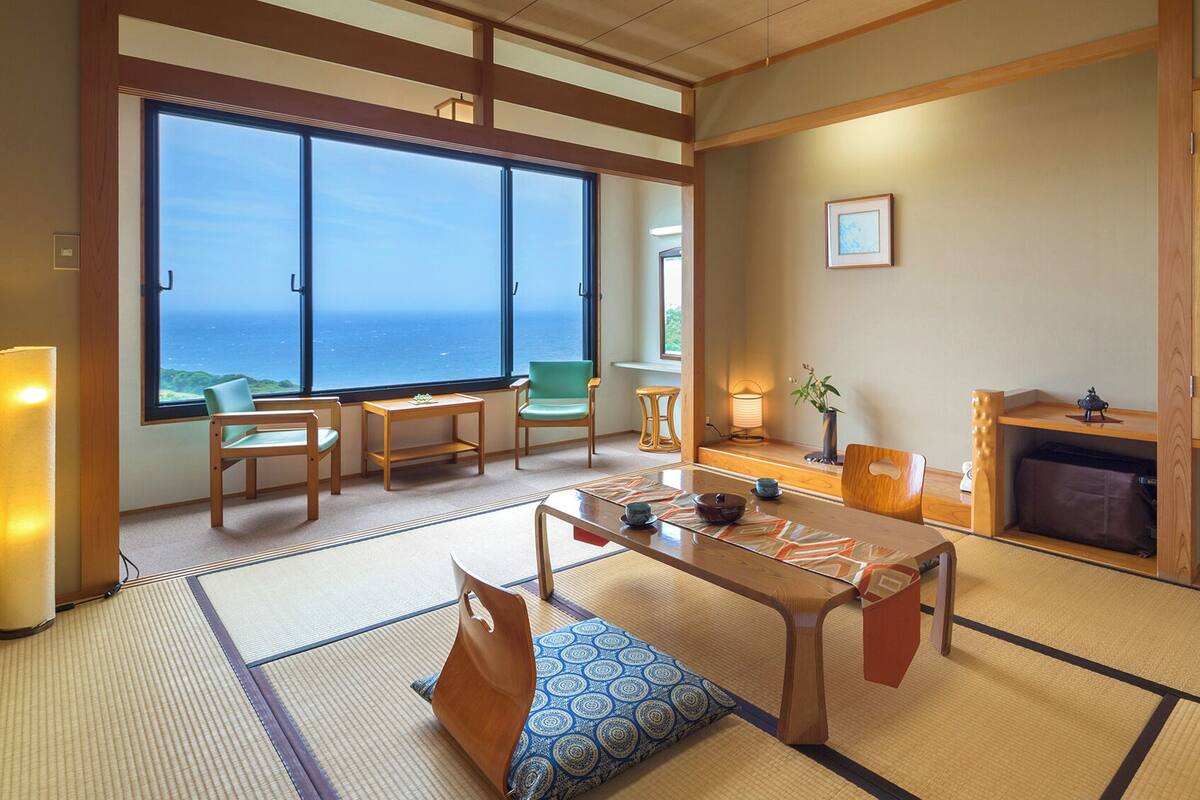 A traditional Japanese room with tatami flooring, a low table, chairs, a view of the sea, and a wooden cabinet.