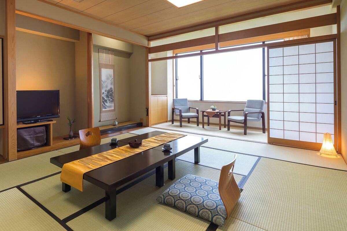 A traditional Japanese room with tatami flooring, a low table, a television, sliding doors, and a view through a large window.