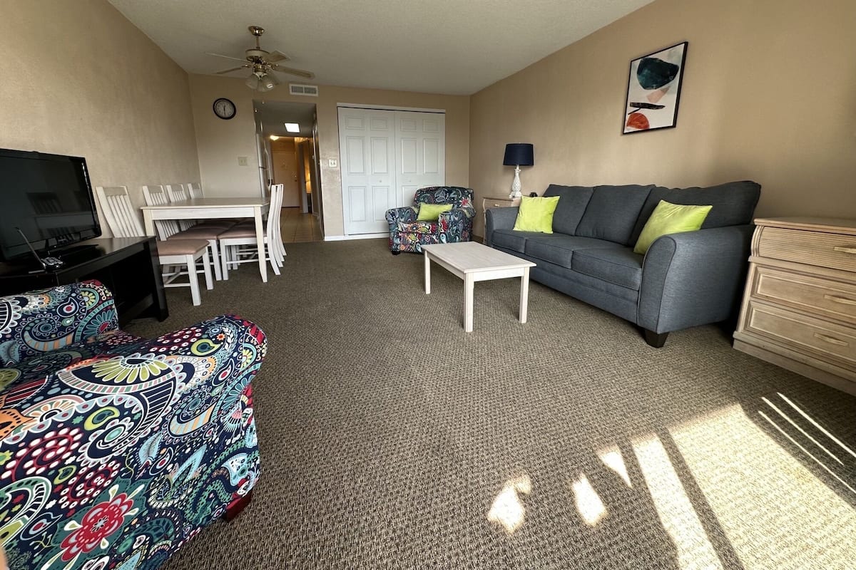 Un salon comprenant un téléviseur, un canapé, une table basse, un pouf à motifs et un coin repas avec des chaises et une table.