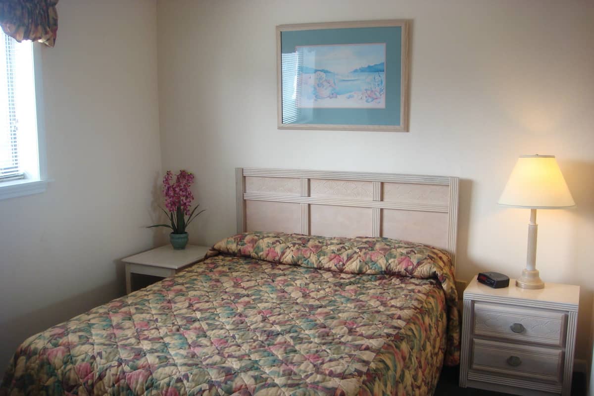 Un lit bien fait, recouvert d’une courtepointe à motifs floraux, une lampe de chevet, une table de chevet avec une radio et un tableau encadré au mur.