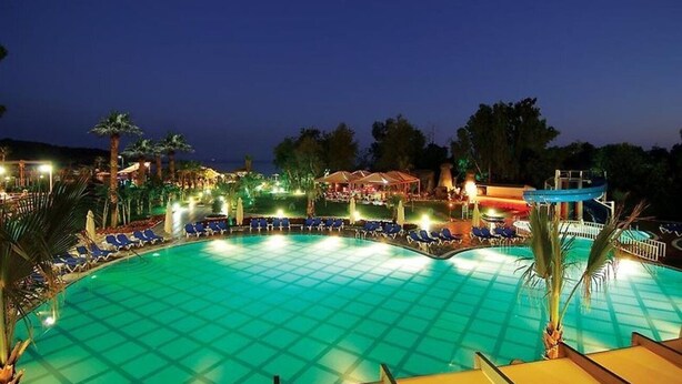 Una gran piscina al aire libre con sillas reclinables azules y palmeras, rodeada de estructuras iluminadas y un cielo nocturno.