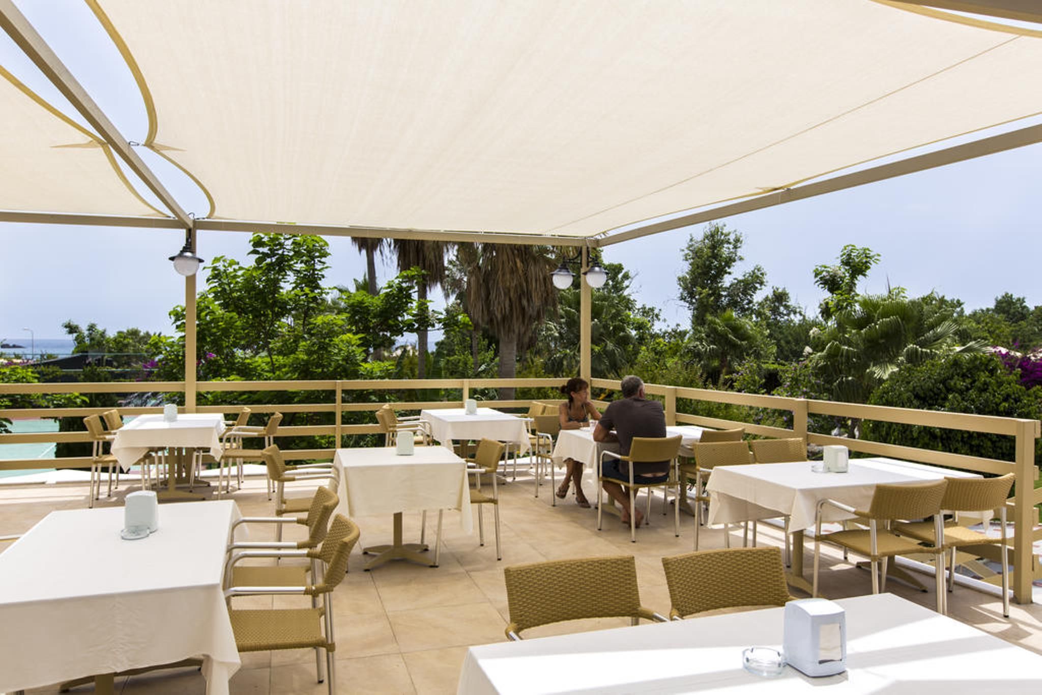 Un espace extérieur aménagé pour les repas, avec des tables et des chaises, une piscine en arrière-plan et une vue sur l’océan.