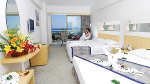 Habitación de hotel con dos camas, un escritorio con un arreglo floral, una silla y un balcón con vista al mar.