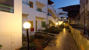 A narrow pathway flanked by white buildings with balconies, a stone wall on one side, and a garden with pink flowers.