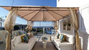 A covered outdoor seating area with two white sofas, a coffee table, and a view of the pool and ocean.