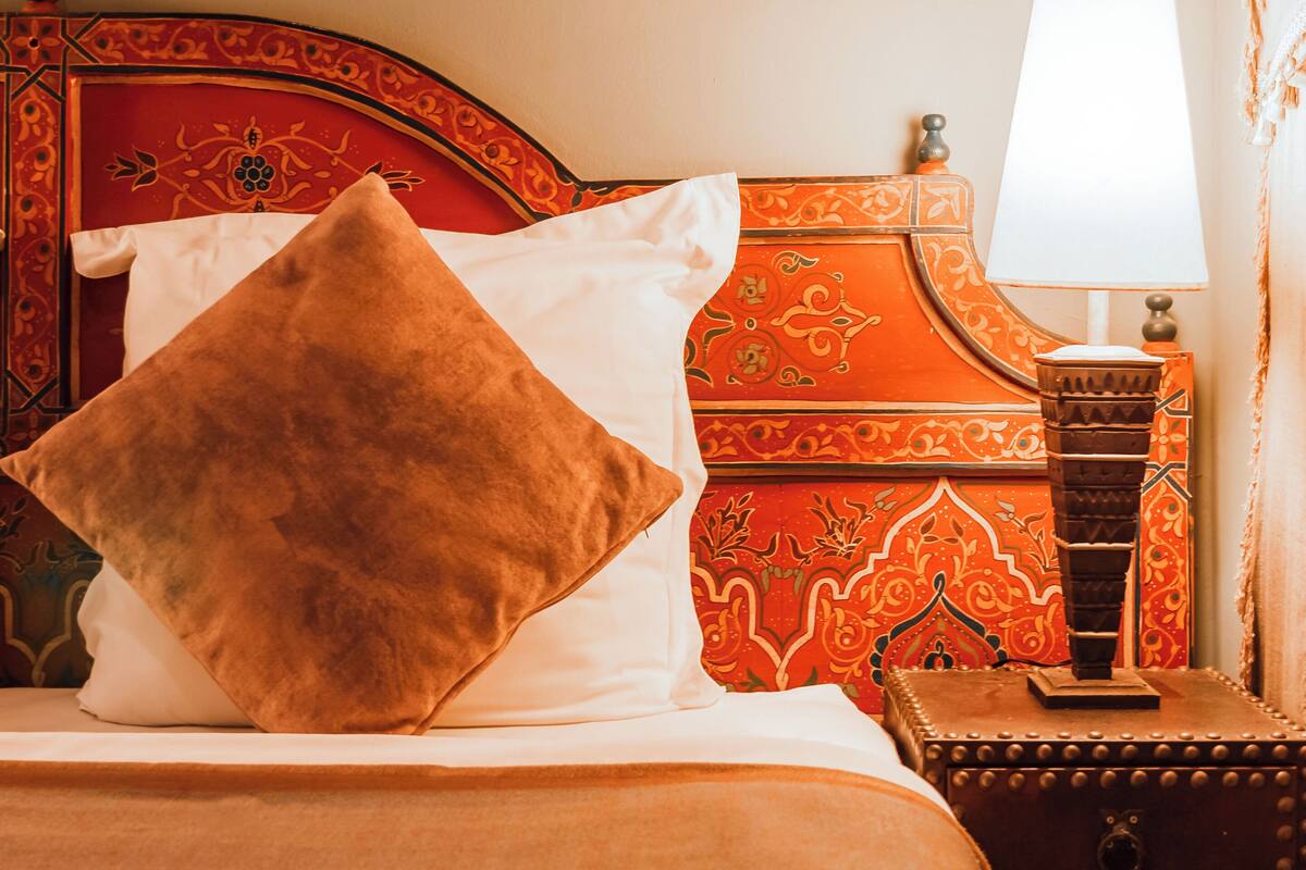 A bed with a red headboard, a white pillow, and a brown pillow. There is a lamp on a nightstand beside the bed.