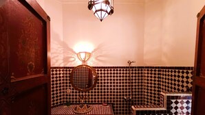 A bathroom with a checkered tile backsplash, a round mirror, a sink with a decorative basin, and a hanging light fixture. - Riad Zagouda (Marrakech)