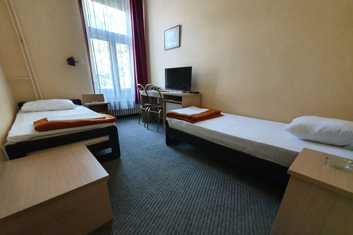 Habitació d'hotel amb dos llits individuals, escriptori, cadira, televisió i finestra amb cortines.