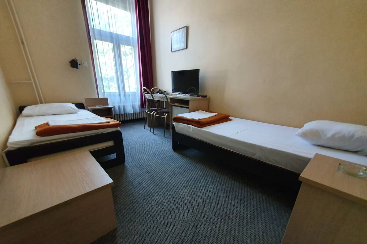 A hotel room with two single beds, a desk, a chair, a TV, and a window with curtains.