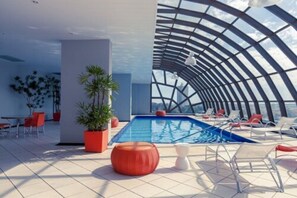Un área de piscina interior moderna con techo curvo, sillones de descanso y plantas en macetas.