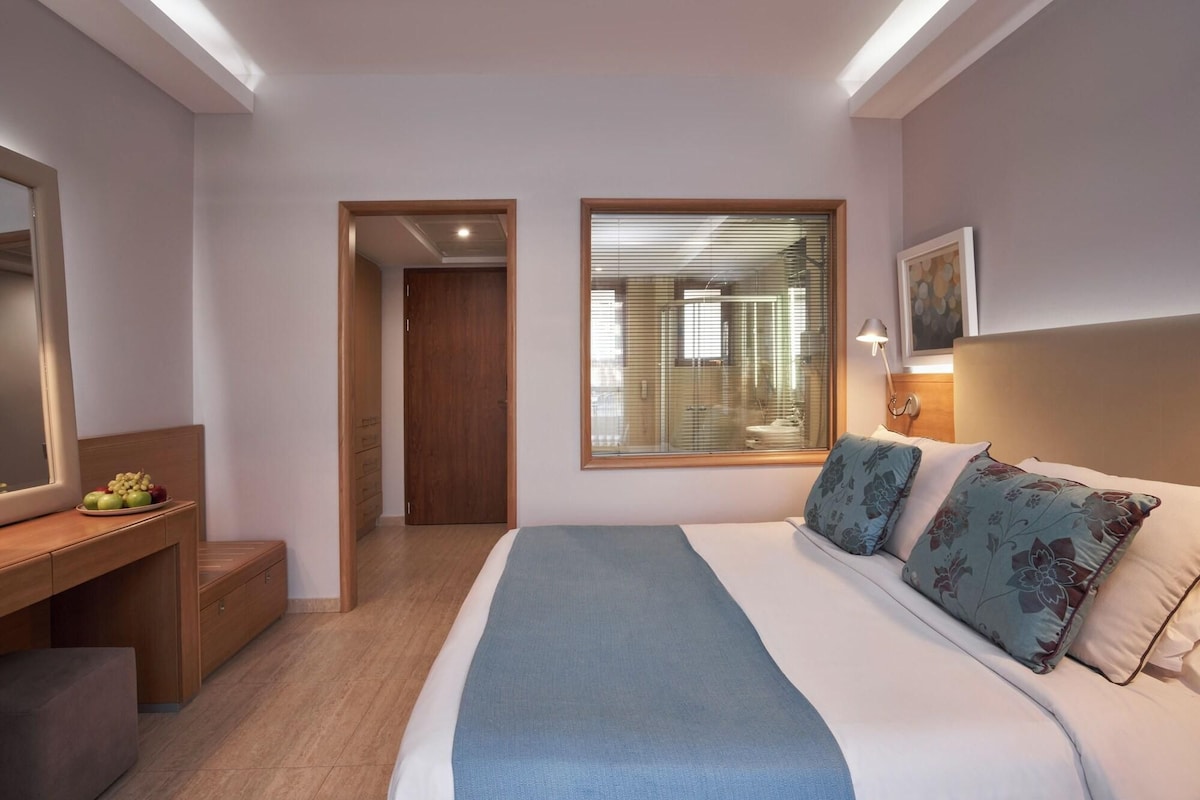 A hotel room with a bed, bedside table, mirror, and a window with blinds.