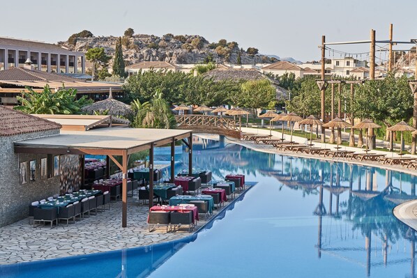 Un espace aménagé au bord de la piscine, avec des tables et des parasols, un coin salon couvert et une vue sur une colline parsemée de bâtiments.