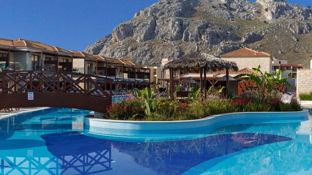 A pool area with a mountain backdrop, a thatched structure, and a view of hotel buildings. - Atlantica Holiday Village Rhodes (Rhodes)