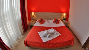 Habitación de hotel con una cama curvada, ropa de cama roja, dos almohadas, una mesita de noche con lámpara y una ventana con cortinas translúcidas.