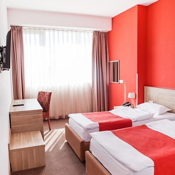A hotel room with two beds, a desk, a chair, and a window with curtains. - Putnik Hotel (Novi Sad)