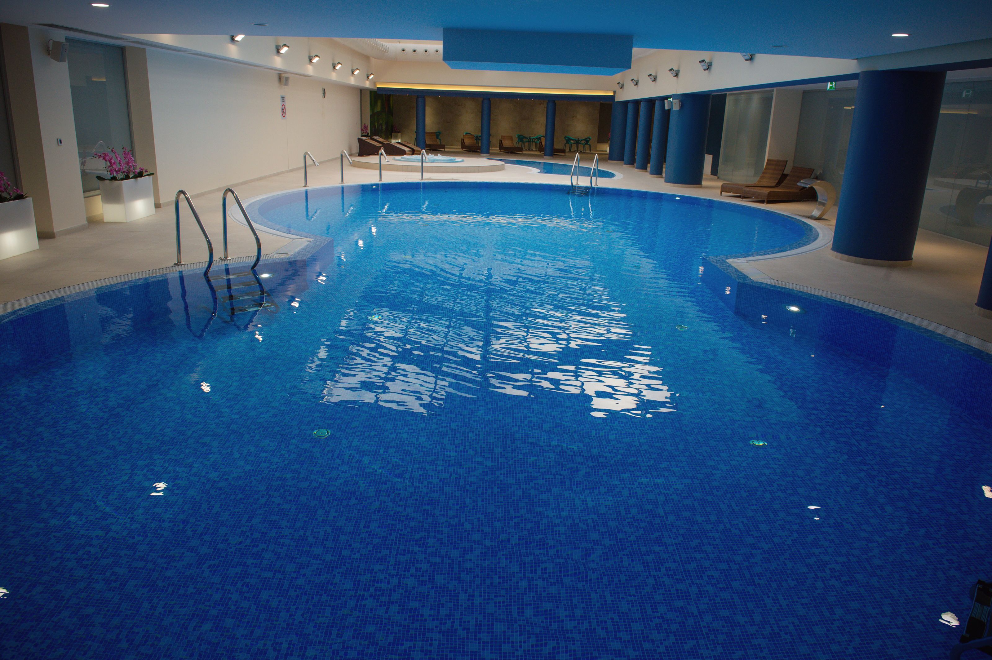 Ein Hallenbad mit klarem Wasser, mehreren Bahnen und Liegestühlen am Rand.
