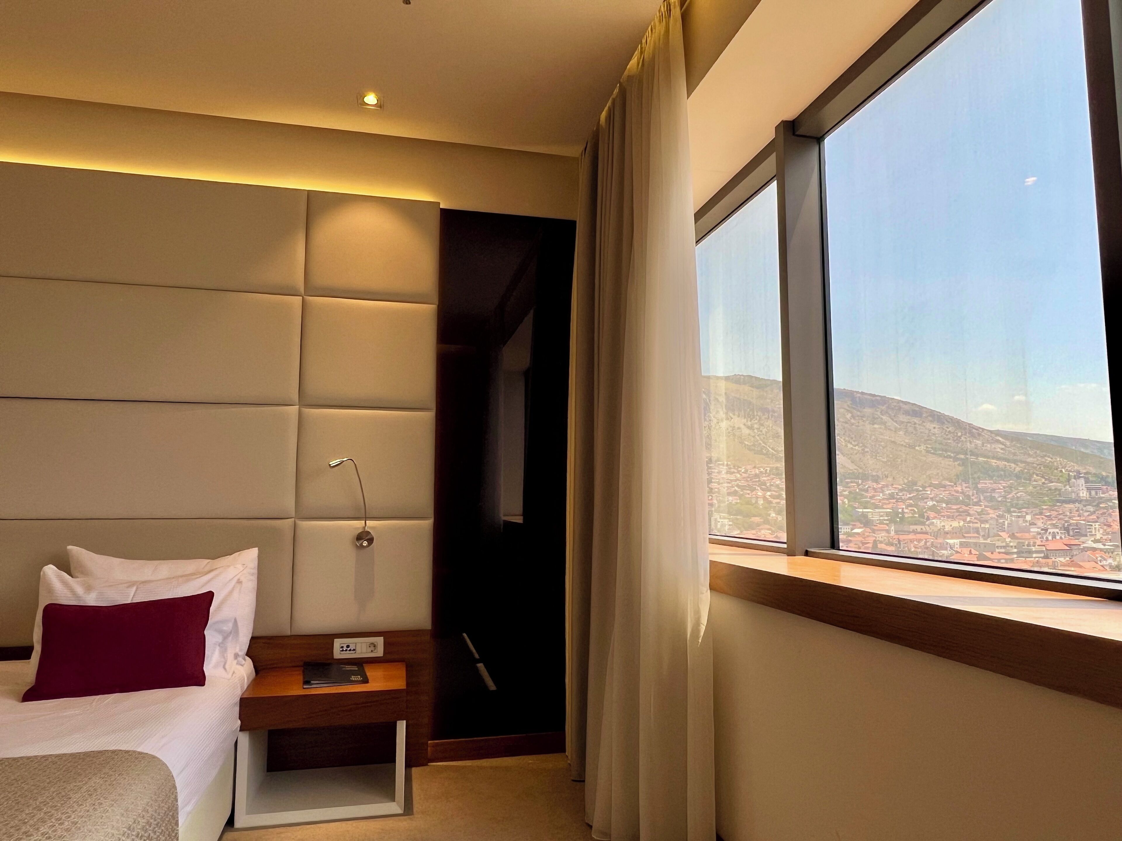 A modern hotel room with a large window offering a city view, a bed with a red pillow, a bedside table with a lamp, and a wall-mounted control panel.