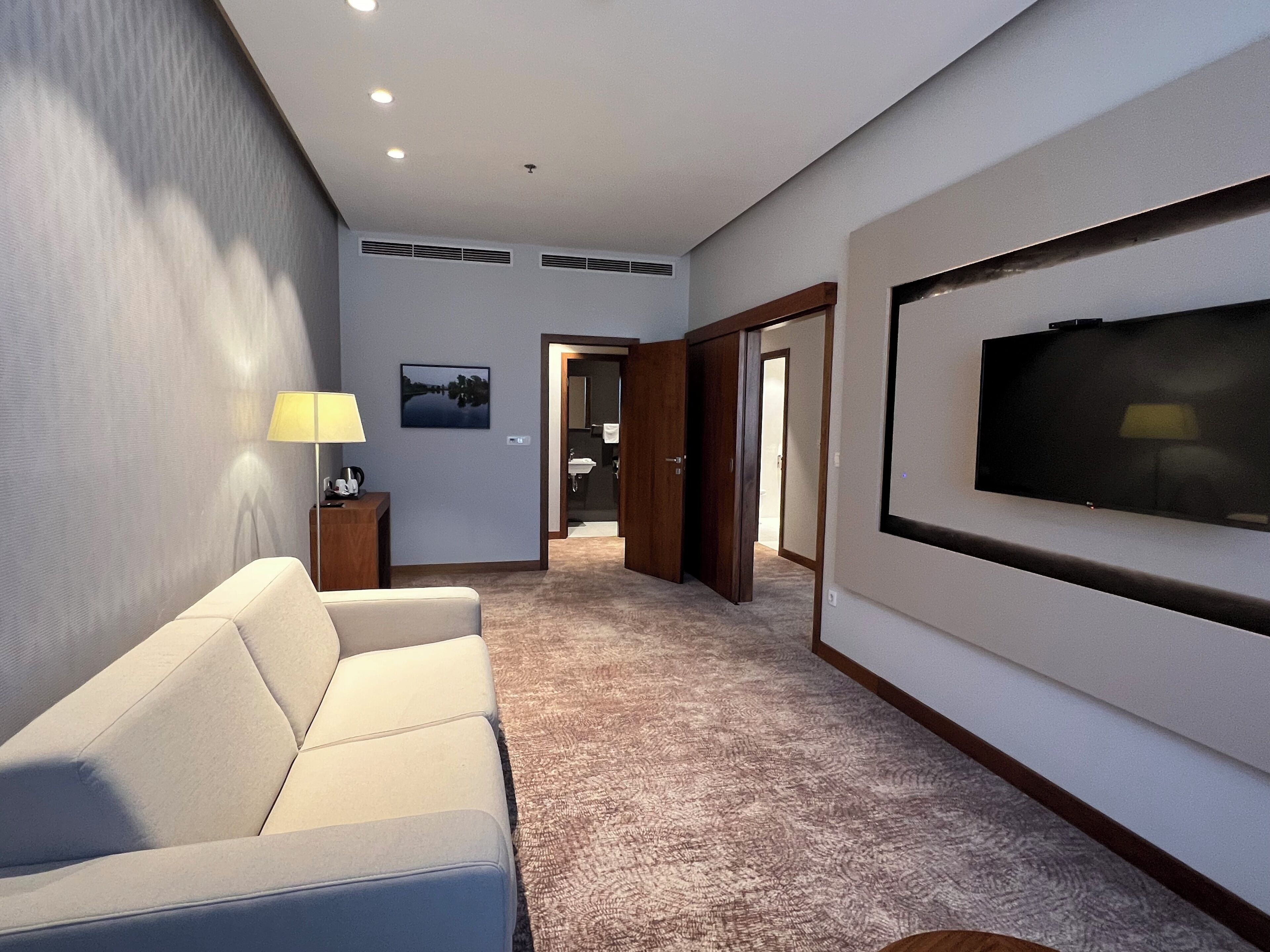 A hotel room with a beige sofa, a flat-screen TV mounted on the wall, a lamp, and a picture of a landscape.