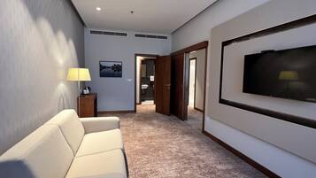 A hotel room with a beige sofa, a flat-screen TV mounted on the wall, a lamp, and a picture of a landscape.