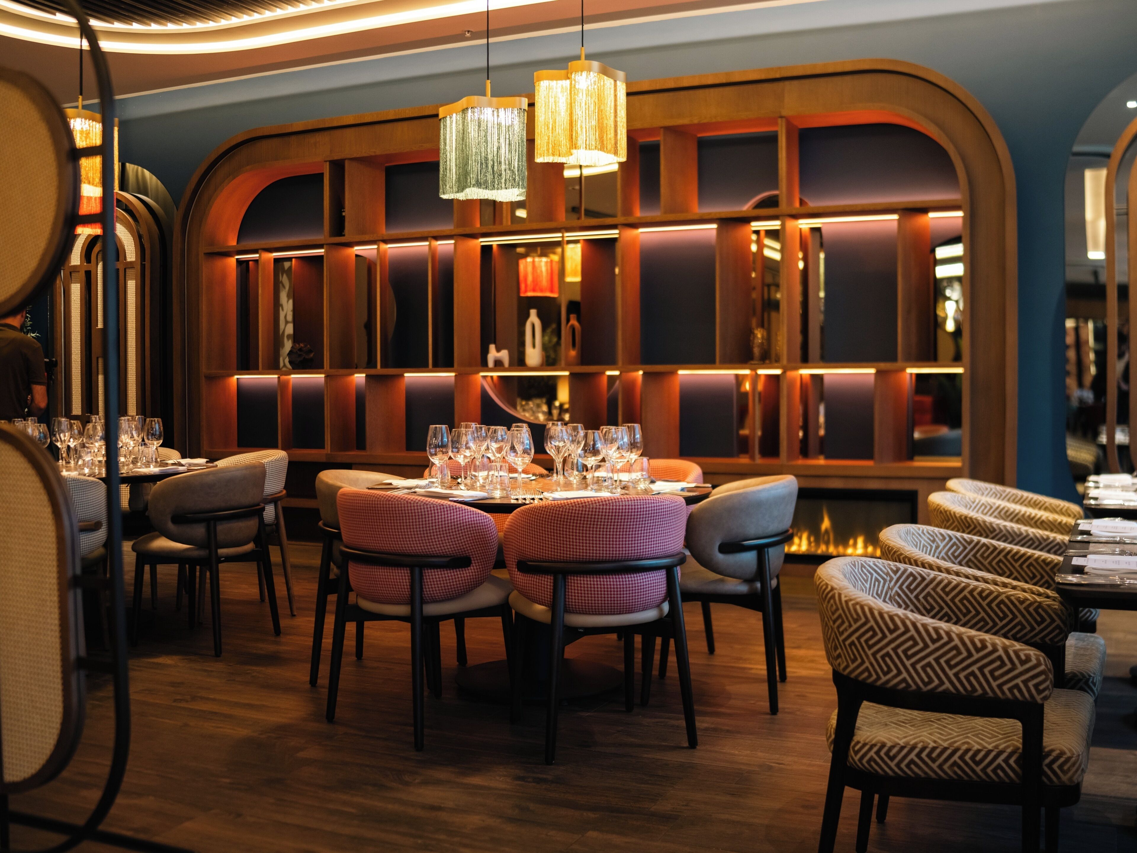 A restaurant interior with a long table set for dining, surrounded by chairs, and a decorative wall with a fire feature.