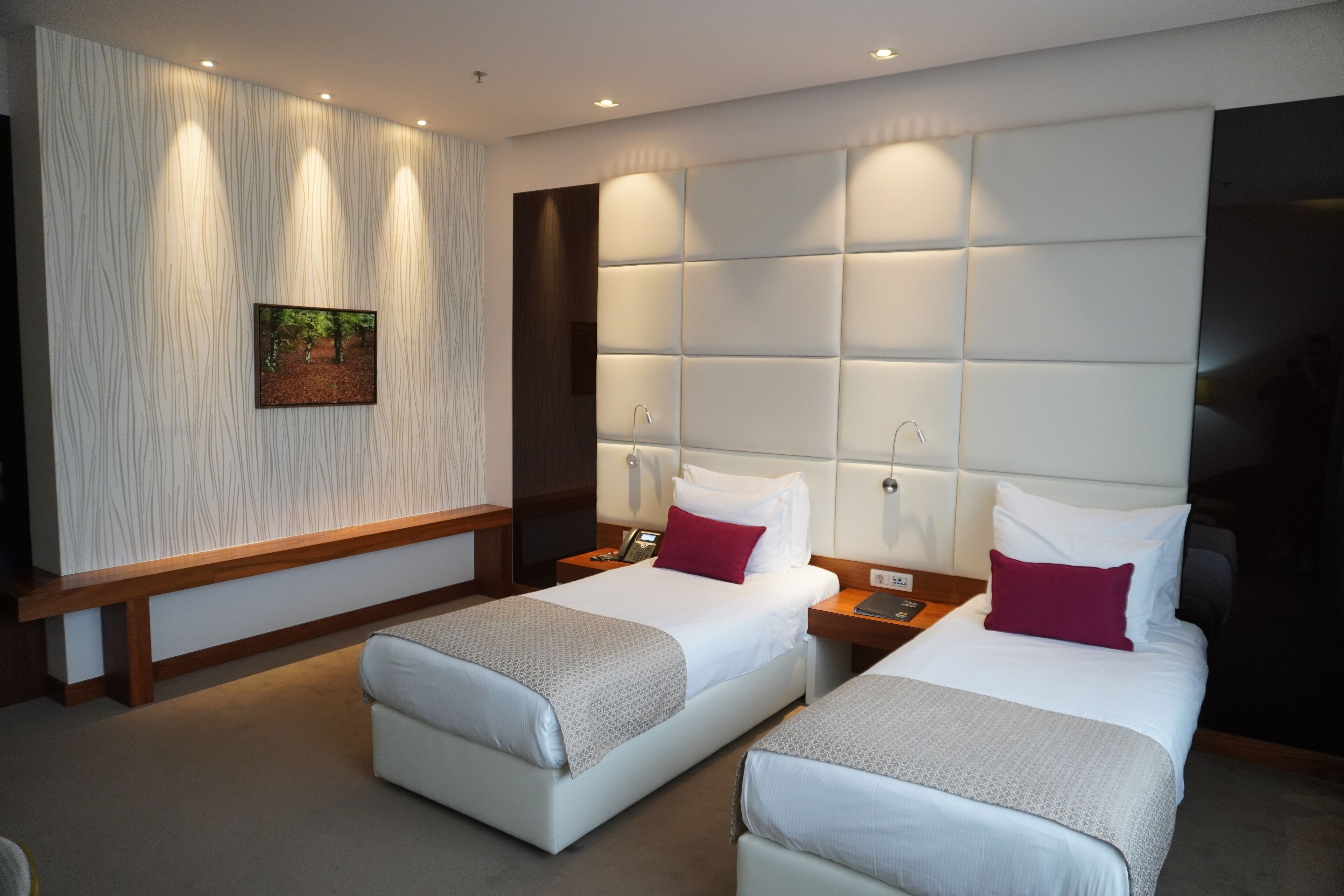 A hotel room with two beds, a large padded headboard, a wooden bench, and a wall-mounted picture.