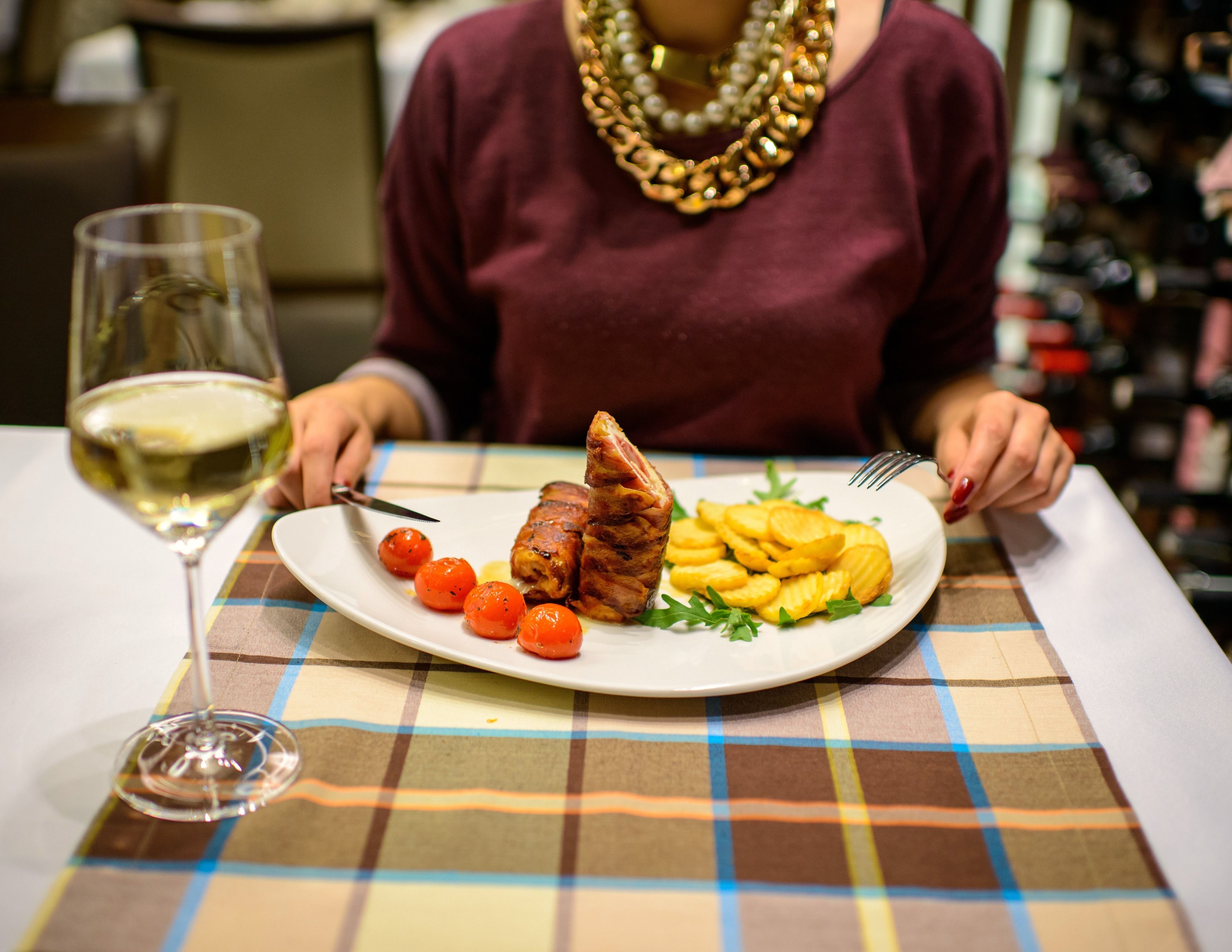 A dining table with a checkered tablecloth, a plate of food, a glass of white wine, and a person wearing a necklace.