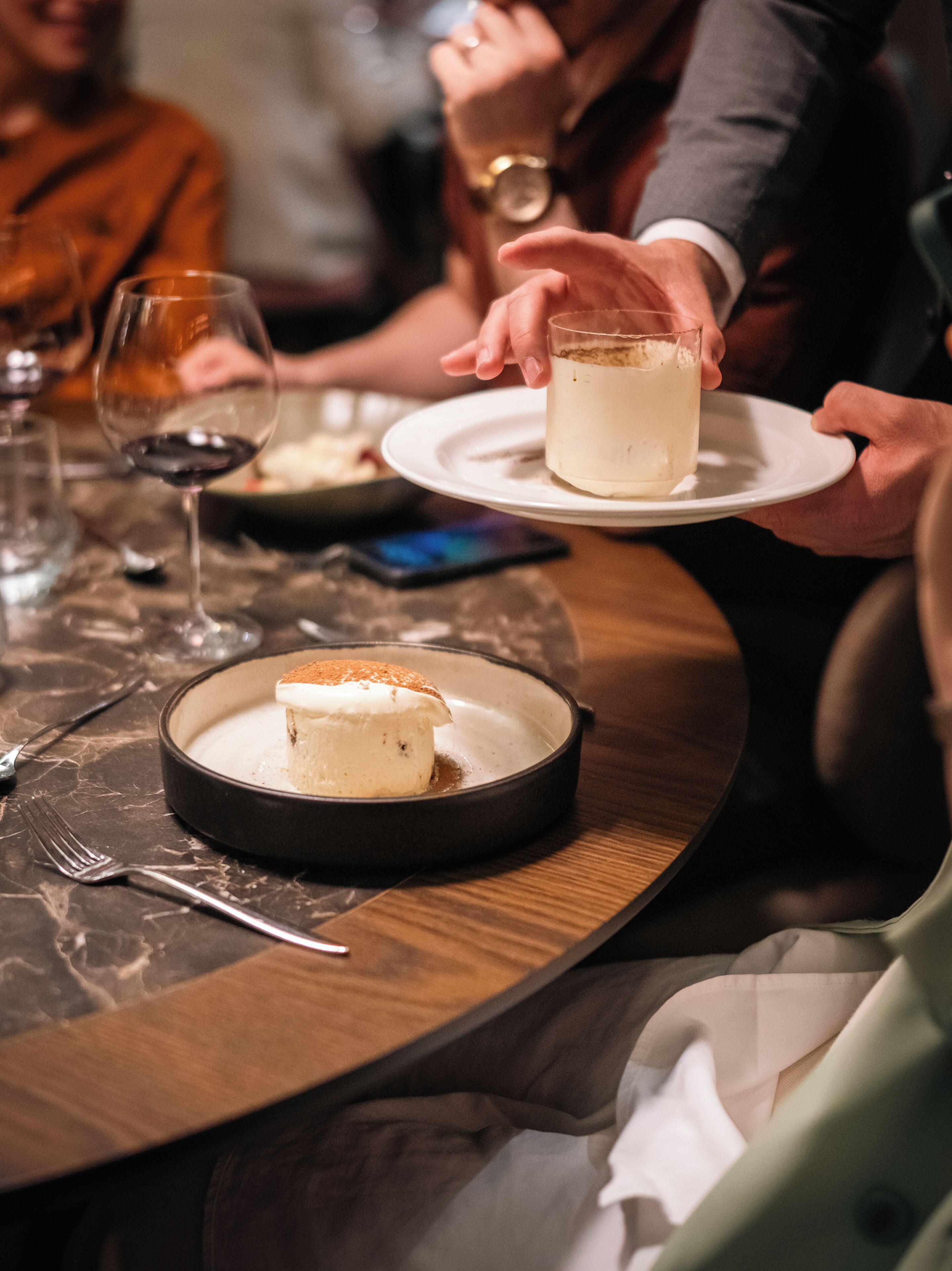 A dining table with a dessert plate, a glass of a light-colored beverage, and a smartphone on the table.