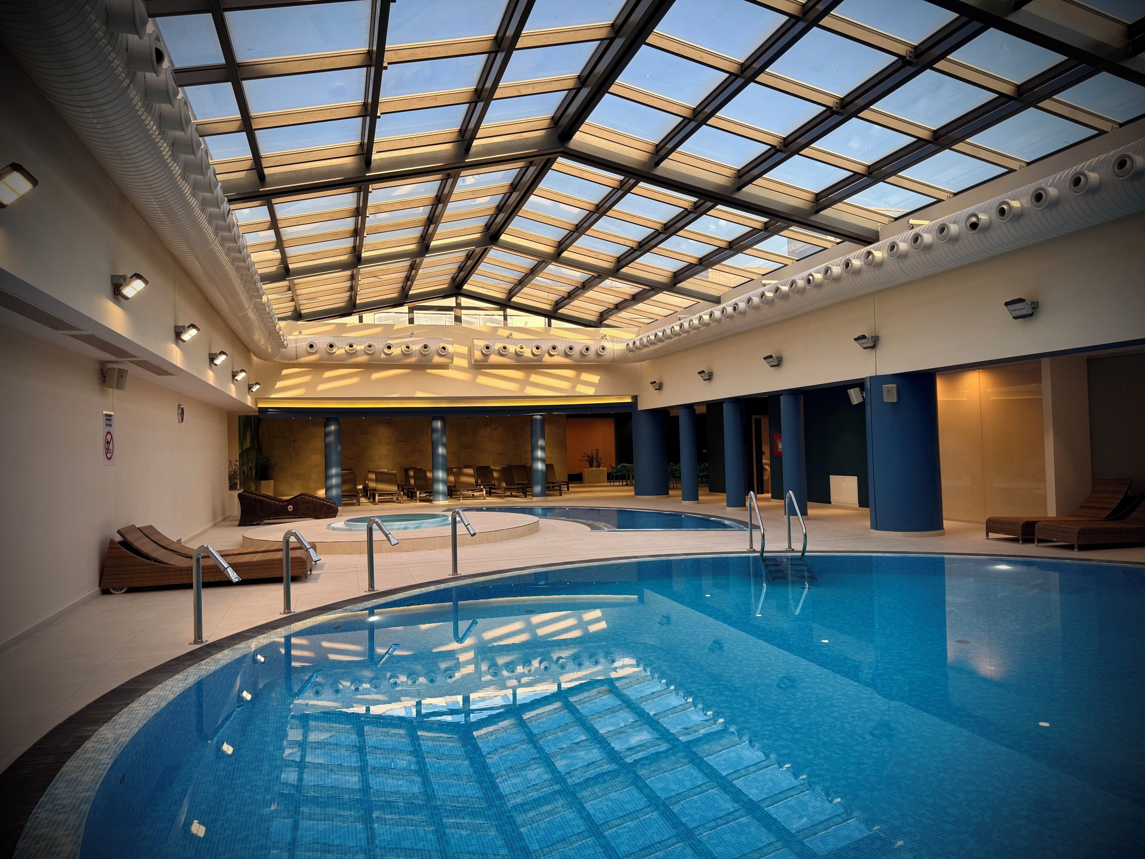 An indoor swimming pool with a glass ceiling, lounge chairs, and a sunlit interior.