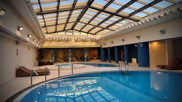 An indoor swimming pool with a glass ceiling, lounge chairs, and a sunlit interior.