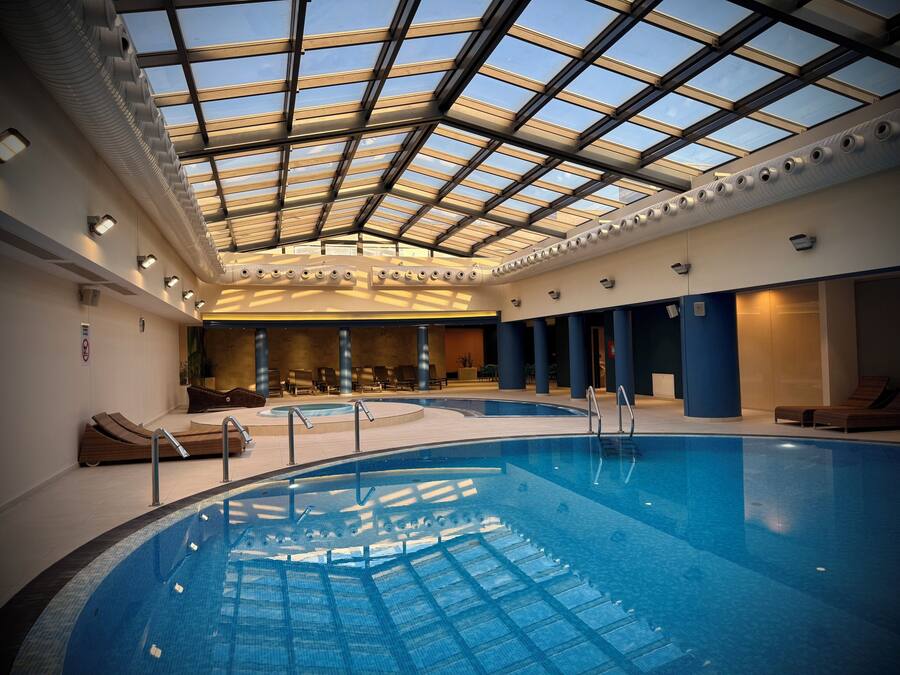 An indoor swimming pool with a glass ceiling, lounge chairs, and a sunlit interior.
