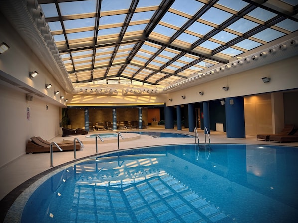 Una piscina interior con techo de cristal, sillas de descanso y un ambiente luminoso.