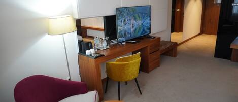 A modern hotel room with a wooden desk, a flat-screen TV, a red armchair, a yellow chair, and a small wooden side table.