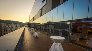 Une terrasse sur le toit, avec des tables et des chaises rondes, une vue sur le paysage urbain et un bâtiment aux grandes fenêtres qui reflètent le coucher de soleil.