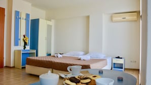 A hotel room with a bed, bedside table with a plate of cookies and a cup, a small table with a teapot and cups, a nightstand with a lamp, and a wall-mounted air conditioner. - Rainbow 1 Holiday Complex (Sunny Beach)